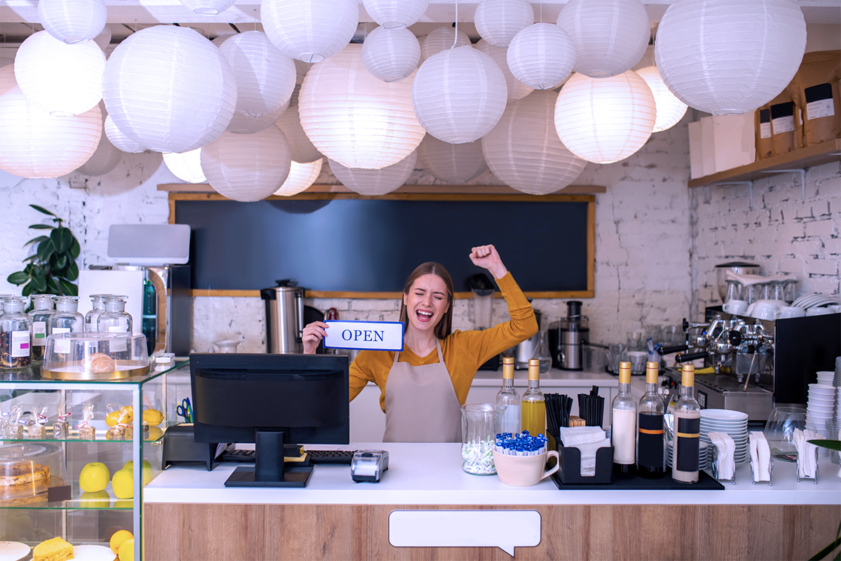 New Small Business Cafe Owner Holding an Open Sign at the Counter Next to Coffee and Cake Displays