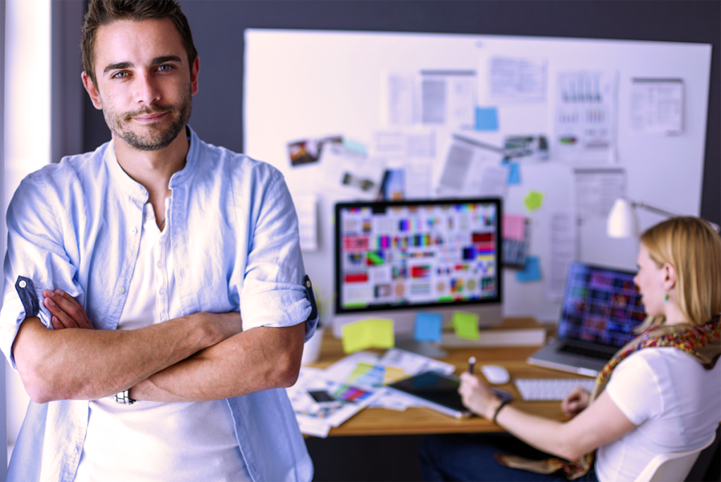 Small Business Owners Discussing Marketing Challenges as the Man is Leaning on an Office Wall with a Woman Sitting at a Desk Crunching Numbers