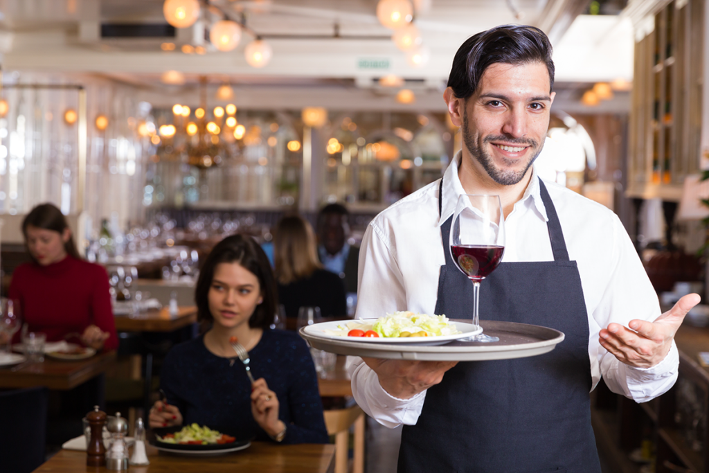 Smiling Waiter Holding a Platter with Salad and a Glass of Red Wine at an Upscale Restaurant Cafe Filled with Customers Eating and Talking and Enjoying Their Atmosphere and Meals