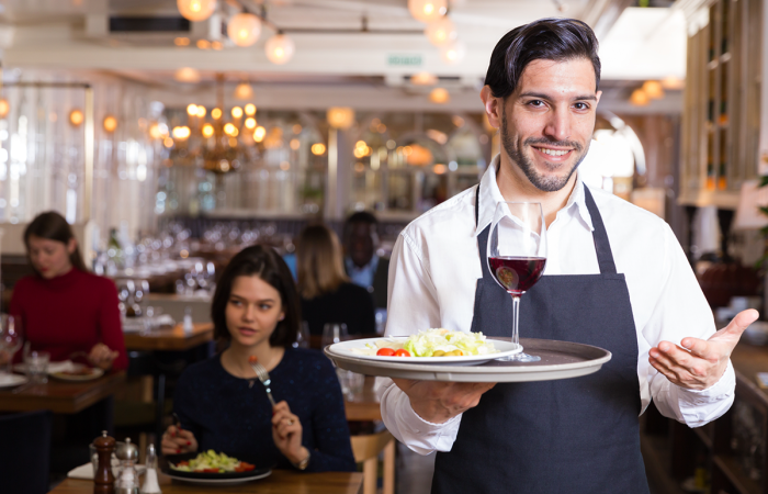 Smiling Waiter Holding a Platter with Salad and a Glass of Red Wine at an Upscale Restaurant Cafe Filled with Customers Eating and Talking and Enjoying Their Atmosphere and Meals