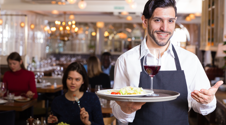 Smiling Waiter Holding a Platter with Salad and a Glass of Red Wine at an Upscale Restaurant Cafe Filled with Customers Eating and Talking and Enjoying Their Atmosphere and Meals